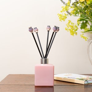 Pode incluir: Frasco difusor de vidro rosa com varetas pretas coroadas com cápsulas de sementes de papoula roxas. O difusor está sobre uma mesa de madeira com um livro e um jarro de vidro com flores no fundo.