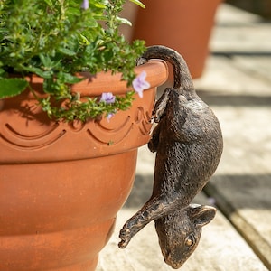 Könnte beinhalten: Ein brauner Keramiktopf mit grünen Pflanzen und lila Blüten. Eine kleine, dunkelbraune, metallene Mausfigur hängt am Rand des Topfes.