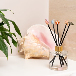 May include: A glass diffuser bottle with a gold lid and black reeds. The reeds are topped with small sea creature figurines, including a starfish, a turtle, a dolphin, and a fish. A large seashell is in the background.