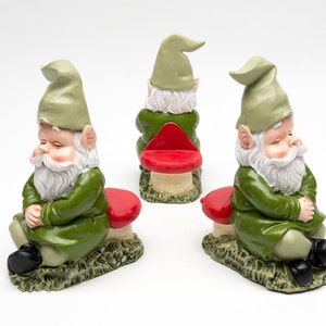 Könnte beinhalten: Drei grüne Gnome mit weißen Bärten sitzen auf roten und weißen Pilzhockern. Die Gnome tragen grüne Hüte und haben die Augen geschlossen.