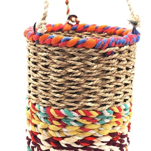 Könnte beinhalten: Ein geflochtener Hängekorb mit einem bunten geflochtenen Seilbesatz. Der Korb ist aus natürlichen Materialien gefertigt und hat einen rustikalen Look.