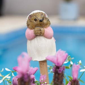 May include: A whimsical garden stake featuring a hedgehog figurine. The hedgehog wears a white bonnet, a pink sweater, and a white dress. The figurine is set against a blurred background of a blue pool and pink flowers.
