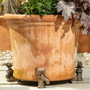 May include: A terracotta planter with a rustic finish sits on four decorative bird-shaped plant stand feet. The feet are made of metal and have a distressed bronze finish.