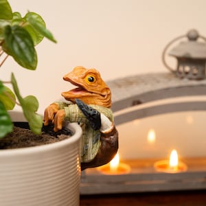 Può includere: Una piccola statuina di rana marrone e gialla con un ampio sorriso, che indossa una camicia a motivi verdi e marroni e una borsa a tracolla marrone, è appollaiata sul bordo di un vaso di ceramica bianco con una pianta verde che cresce al suo interno. Il vaso è posto su una superficie di legno davanti a un portacandele in metallo grigio con quattro candele accese.