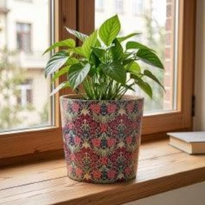 May include: A cylindrical plant pot featuring a repeating damask pattern in shades of deep red, navy blue, and muted green. The pot contains a lush green houseplant with pointed leaves, positioned upright with a smooth, flat rim at the top edge.