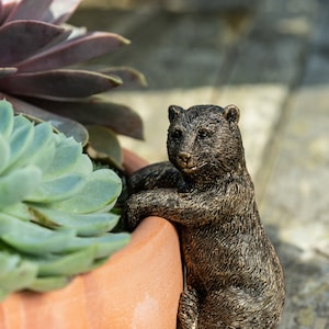 May include: A bronze-colored bear figurine is perched on the edge of a terracotta pot, its paws resting on the rim. The bear has a realistic, detailed sculpt with a textured fur finish. The pot is filled with succulent plants.