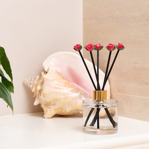 May include: A glass diffuser bottle with a gold lid and black sticks topped with red rose accents. The bottle is filled with clear liquid and is sitting on a white surface next to a large seashell.