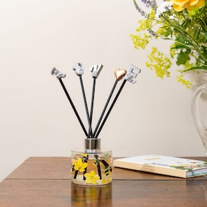 Op de afbeelding: Een glazen diffuserfles met zwarte rietjes en metalen bedels. De fles heeft een geel en zwart bijendesign en gele bloemen. De bedels spellen het woord "LOVE" uit.