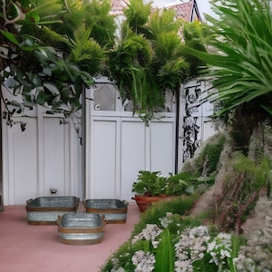 May include: Three galvanized metal planters in different sizes, with two smaller planters in front of a larger planter. The planters are sitting on a pink concrete patio in front of a white wooden fence. The fence is covered in green vines and plants.