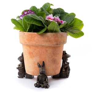 Op de afbeelding: Een terracotta bloempot met een roze en paarse bloeiende plant staat op een set van drie bronzen konijnen plantenvoeten.