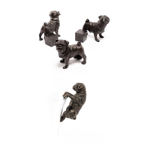 May include: Four bronze pug figurines, three standing on small square bases and one hanging from a glass object. The pugs are all in different poses.