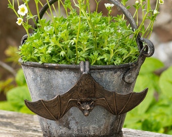 Peut inclure: Un pot de fleurs en métal vieilli avec un ornement de chauve-souris en bronze. Le pot contient des plantes vertes et de petites fleurs blanches. La chauve-souris est centrée sur le devant du pot.