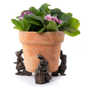 Op de afbeelding: Een terracotta bloempot met een roze en paarse bloeiende plant. De pot staat op drie decoratieve konijnenfiguren.
