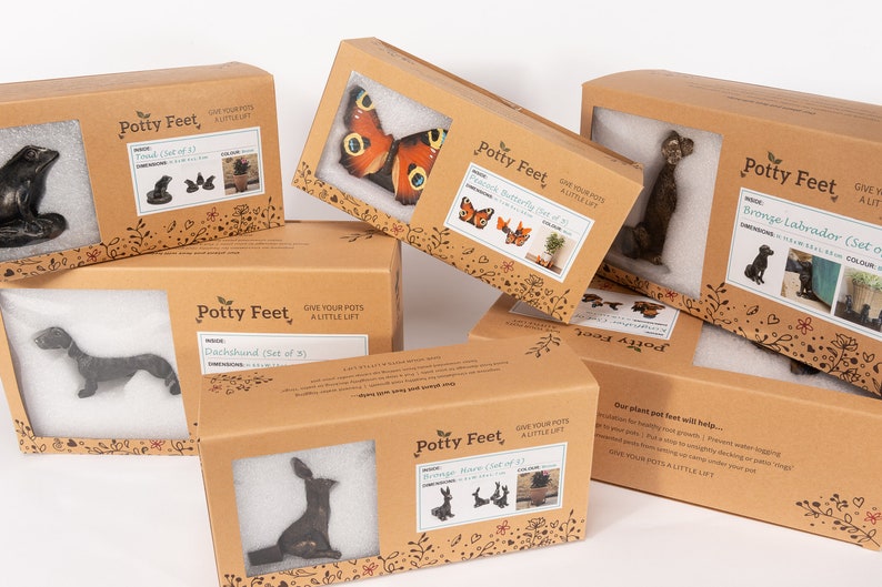 May include: Assortment of cardboard boxes with the text "Potty Feet" printed on them. The boxes contain various animal and butterfly figurines, designed to be used as plant pot feet. The boxes are stacked on top of each other, with the text "Give your pots a little lift" printed on the front of each box.
