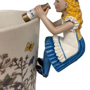 May include: A figurine of a girl with blonde hair, wearing a blue dress and white apron, perched on the edge of a floral-decorated mug. She holds a small bottle with the text "K ME".