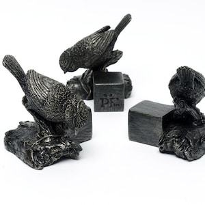May include: Three silver-toned bird sculptures. Each bird is perched on a textured base, with one on a small cube. The sculptures have a weathered appearance. The background is white.