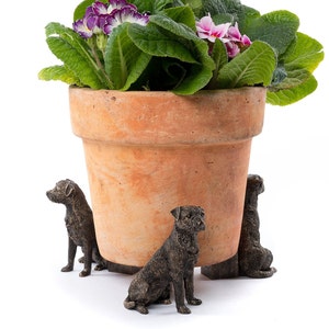 Puede incluir: Tres pequeñas figuritas de perros de bronce están colocadas alrededor de la base de una maceta de terracota. La maceta está llena de una planta verde frondosa con flores moradas y rosas.