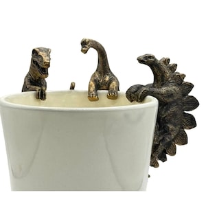 Könnte beinhalten: Ein weißer Keramik-Pflanztopf mit drei bronzefarbenen Dinosaurierfiguren am Rand. Die Dinosaurier umfassen einen T-Rex, einen Brontosaurus und einen Stegosaurus. Der Topf steht vor einem weißen Hintergrund.