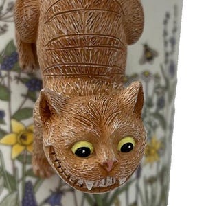 Könnte beinhalten: Eine Keramiktasse mit einem 3D-Design der Grinsekatze. Die Katze ist orange mit einem breiten Grinsen und gelben Augen. Die Tasse hat ein Blumenmuster mit gelben und lila Blumen.