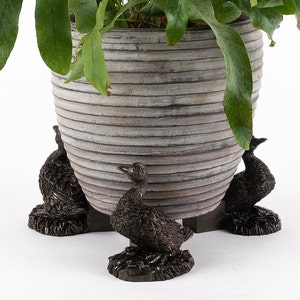 May include: A grey ceramic planter with a ribbed design sits on three decorative duck-shaped plant stands. The planter is filled with a green leafy plant.