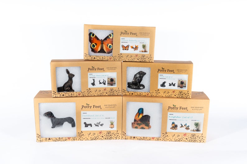 Puede incluir: Seis cajas diferentes de pies para macetas, cada una con un dise&ntilde;o de animal diferente. Las cajas est&aacute;n apiladas en forma de pir&aacute;mide. Los animales incluyen una mariposa, un conejo, una foca, un dachshund, un mart&iacute;n pescador y una mariposa. Las cajas son todas marrones y tienen las palabras "Potty Feet" impresas en ellas.