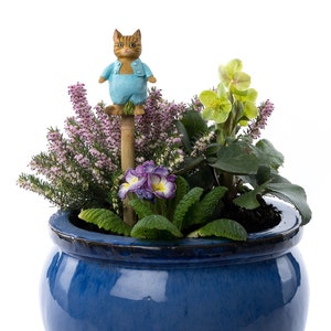 May include: A blue ceramic pot containing a variety of plants, including pink heather, green leaves, and purple flowers. A small figurine of a cat wearing blue overalls is perched on a wooden stick in the centre of the pot.
