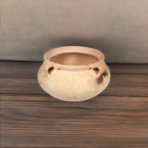 May include: A small, round, terracotta pot with a light brown glaze. The pot has three small, decorative handles on the rim.