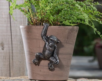 May include: A brown ceramic planter with a monkey figurine hanging on the side. The monkey is black and has a textured finish. The planter is filled with green ferns.