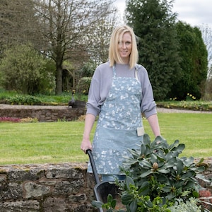 May include: A woman wearing a grey sweater and a blue floral apron is pushing a wheelbarrow full of a green plant. She is standing in a garden with a stone wall and green grass.