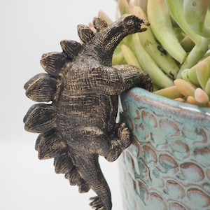 Op de afbeelding: Een gouden dinosaurusfiguur met een stekelige rug, die aan de zijkant van een groene en blauwe keramische pot klimt.