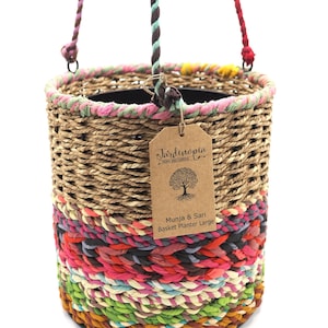 May include: A large hanging basket planter made of woven natural fibres with a colourful striped design. The basket has a brown tag that reads "Jardinopia Home and Garden Munja & Sari Basket Planter Large".
