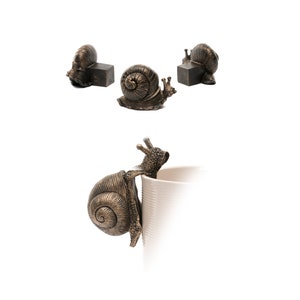 Könnte beinhalten: Drei bronzene Schneckenfiguren, eine hält sich an einer weißen Tasse fest. Die Schnecken haben detaillierte Schalen und sind ein dekorativer Akzent.