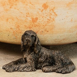 May include: A bronze figurine of a brown dog lying down. The dog has a long, furry coat and is looking at the camera.