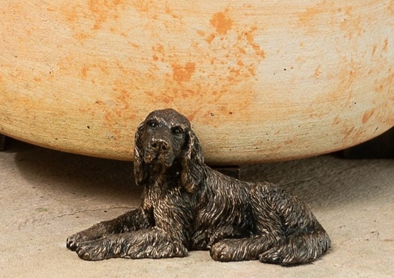 May include: A bronze figurine of a brown dog lying down. The dog has a long, furry coat and is looking at the camera.