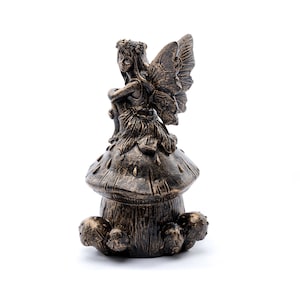 Pode incluir: Uma figura de fada de bronze sentada em uma base em forma de cogumelo. A fada tem asas e uma coroa de flores. O cogumelo tem uma superfície texturizada.