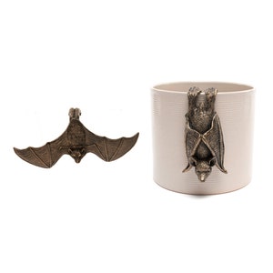 May include: Bronze-coloured bat-shaped decorative items. One bat is shown with its wings spread, and the other is attached to a cream-coloured cylindrical container. The bat has detailed wings and a small face.