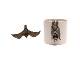 May include: Bronze-colored bat-shaped decorative items. One bat is shown with its wings spread, and the other is attached to a cream-colored cylindrical container. The bat has detailed wings and a small face.