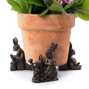 Op de afbeelding: Een terracotta bloempot met een groene plant en roze bloemen staat op een set van drie bronzen konijnen plantenvoeten. De konijnenvoeten hebben een gieter en een konijn dat een bloem vasthoudt.