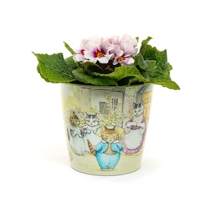 May include: A pink and white primrose plant in a ceramic pot with a colourful illustration of cats. The pot features a scene of cats in a garden setting.