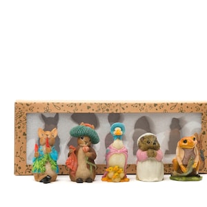 May include: A set of five small figurines in a brown box with a white background. The figurines are of Peter Rabbit, Benjamin Bunny, Jemima Puddle-Duck, Mrs. Tiggy-Winkle, and a frog. The figurines are all in different poses and are made of painted ceramic.