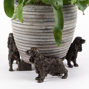 May include: A large, grey, textured plant pot with a green plant, supported by three bronze-coloured dog figurines. The dogs are Cocker Spaniel sculptures, adding a decorative touch to the plant pot.
