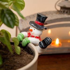 Könnte beinhalten: Eine skurrile Schneemannfigur auf einem Blumentopf. Der Schneemann trägt einen schwarzen Zylinder mit rotem Band, einen roten Schal und grüne Handschuhe. Im Hintergrund sind eine dekorative Laterne und brennende Kerzen zu sehen.