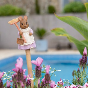 May include: A small ceramic figurine of a rabbit wearing a purple dress and holding a broom. The figurine is standing in a flower pot with pink flowers and green leaves. The background is a blurry image of a swimming pool.