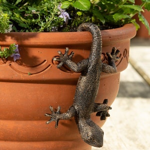 Pode incluir: Uma escultura de lagarto em bronze está pendurada na lateral de um vaso de flores de terracota cheio de vegetação. O lagarto tem escamas detalhadas e está posicionado como se estivesse escalando. O vaso tem uma borda decorativa e é preenchido com várias plantas verdes e pequenas flores roxas.