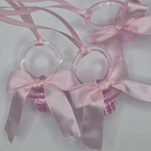 May include: Three pink plastic baby pacifier shaped charms with pink satin ribbon bows attached. The charms are strung on a pink satin ribbon.