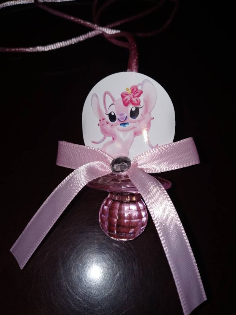 Puede incluir: Collar de chupete rosa y blanco para un baby shower con un personaje de dibujos animados que lleva una flor en la cabeza. El chupete est&aacute; hecho de vidrio transparente con un acabado de purpurina rosa.