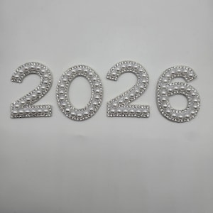 May include: The year 2026 is displayed in a row. Each number is white and outlined with small, silver-colored beads. The numbers are set against a plain white background.