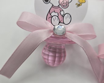 May include: A pink baby pacifier with a Snoopy graphic and a pink ribbon bow. The pacifier has a clear, faceted nipple and a decorative jewel at the bow's center. A small yellow bird is also on the pacifier's face.