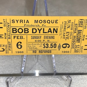 May include: A yellow ticket for a Bob Dylan concert at the Syria Mosque in Pittsburgh, Pennsylvania on February 6, 1966. The ticket is for the first balcony, row M, seat 11 and cost $3.50. The ticket reads "Good Only Sun. Eve. February 6, 1966. Syria Mosque. First Balcony $3.50. Section C Right. Row M 11. Seat"