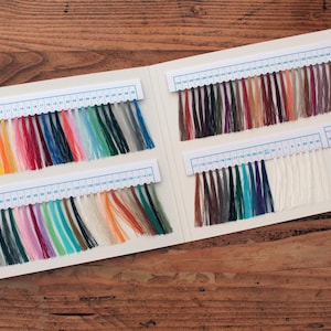 May include: A color chart featuring numerous embroidery thread samples organized in rows on white cards. Each thread is numbered and displays a wide spectrum of colors, including vibrant reds, blues, greens, yellows, and various shades of white and gray.